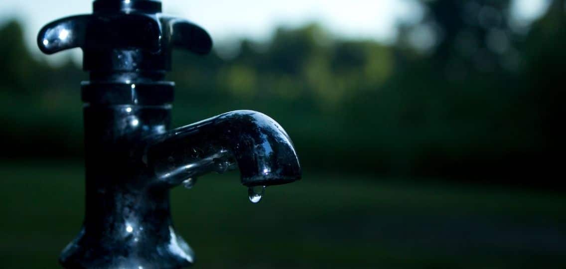 A silver tap outdoors has a single drop of water about to fall from the spout.