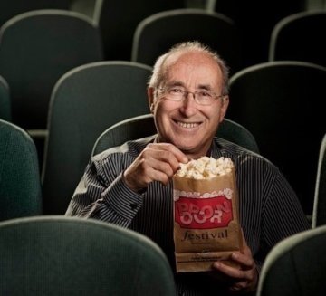 Leonard smiles and sits in a theater with popcorn. He wears glasses and has short, grey hair.