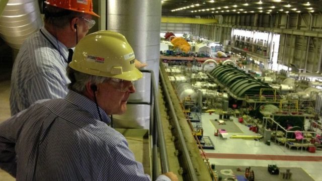 Two workers in hard hats look over a nuclear plant floor below.