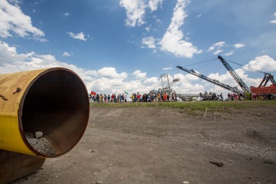A large pipe sits on the sandy ground. An oil rig is in the background and a large group of people gather near it.