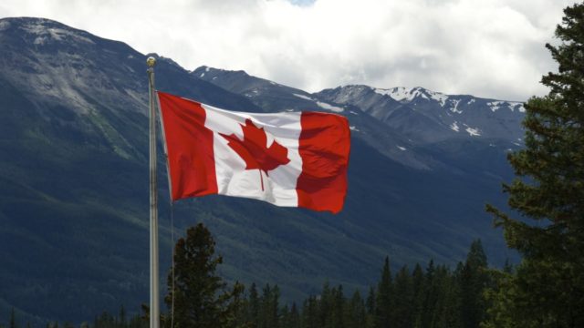 A Canadian flag flies on a pole. Snow capped mountains stand in the background.