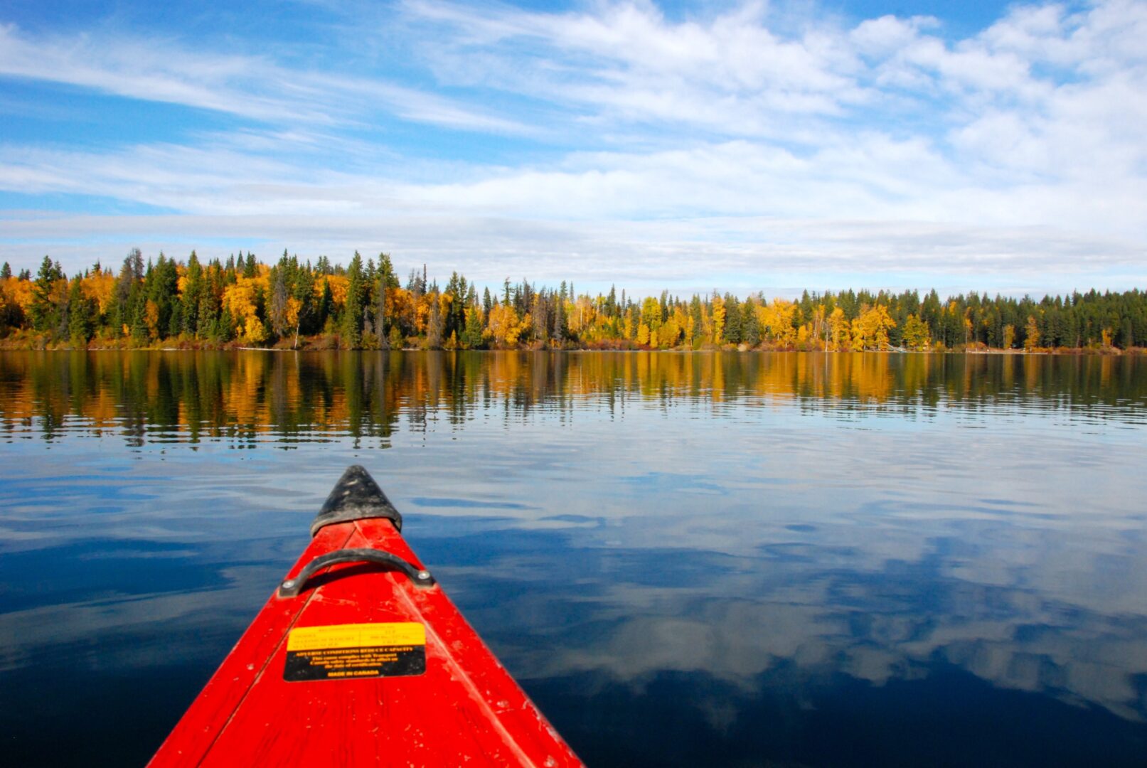 The nose of a red kayak wades out into blue water. Trees with autumn colours stand on the distant shore.