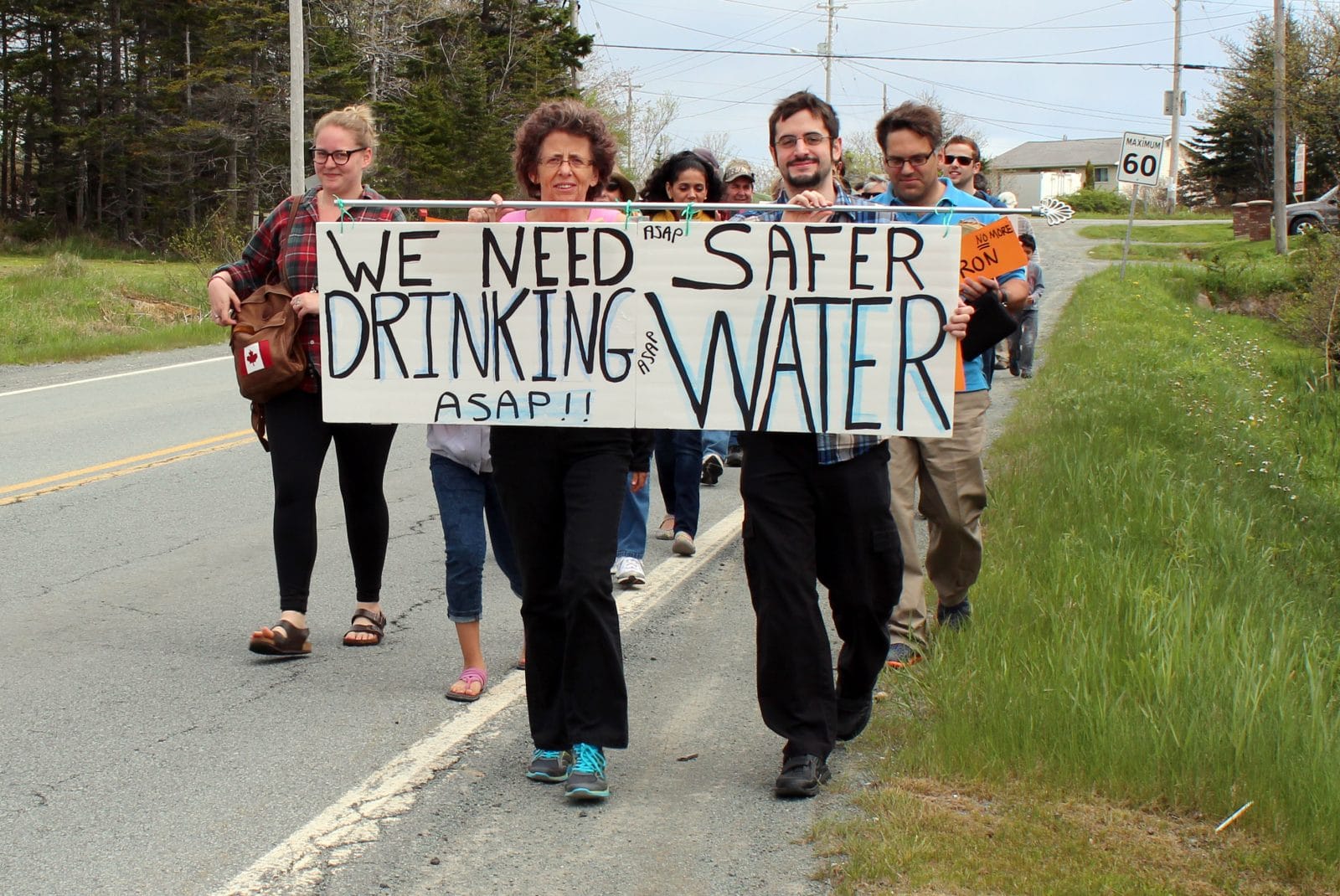 Protesters march along a road. 2 hold a banner that reads "We need safer drinking water ASAP"