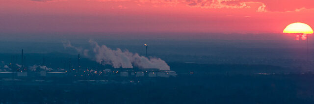 An aerial view shows an industrial complex. Smoke pours into the air around the complex. The sun sets in the distance making the sky pink.