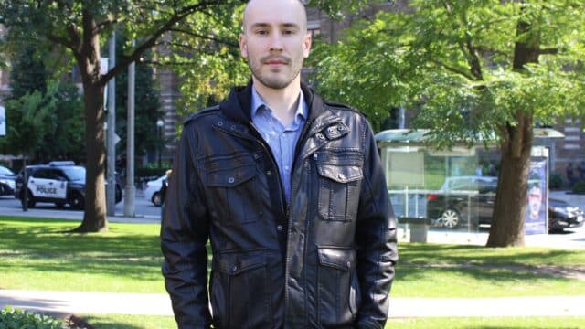 Ian wears a black leather jacket standing in front of trees with a city street in the background.