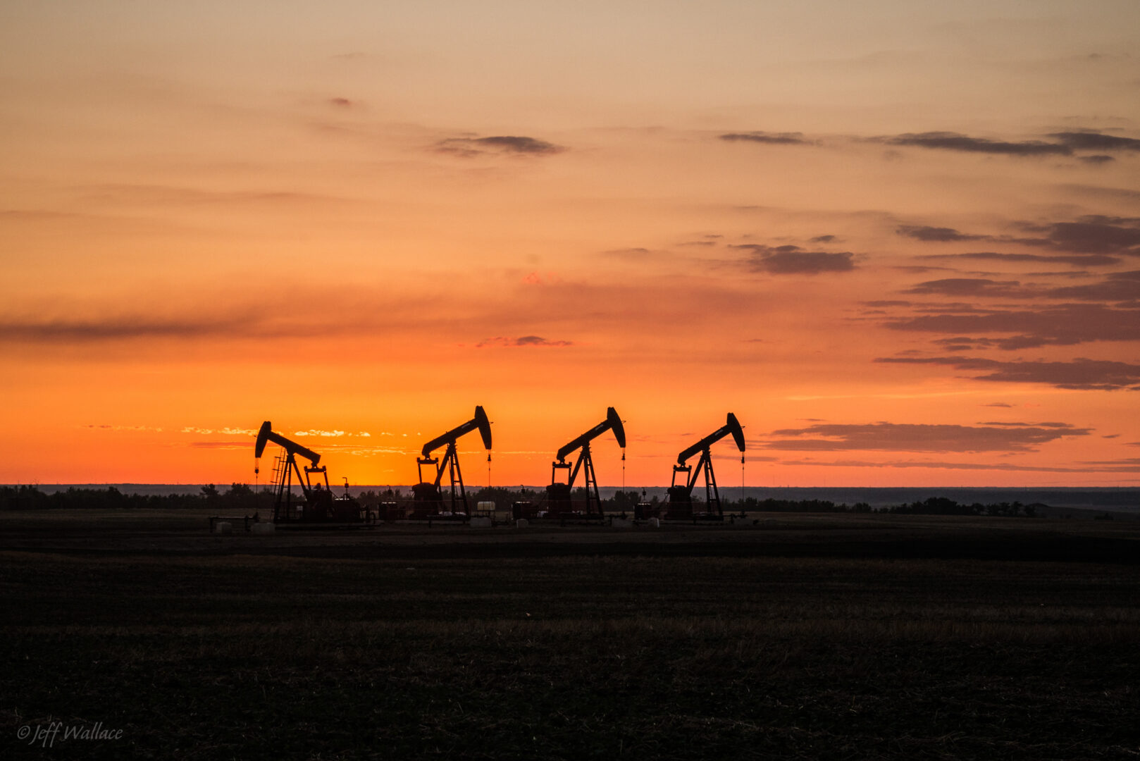 4 oil pumps are placed on the dark ground. The sun sets in the background silhouetting the pumps.