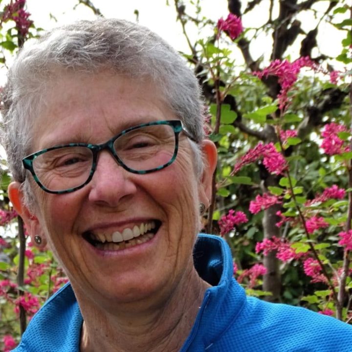 Lynda smiles at the camera. She is outside standing in front of flowers. She wears blue and black glasses, a blue shirt, and has short, white hair.