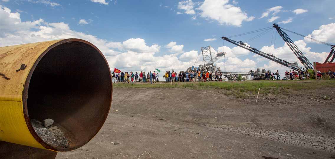A large pipe is placed in the dirt on a flat field. In the distance, a gathering of people stand next to an oil pump.