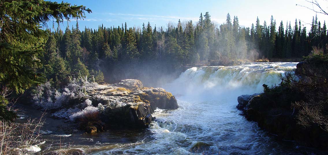 A waterfall creates mist around a rocky cliff. Evergreen trees stand in the background.