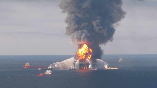 An oil rig is on fire in the middle of the ocean. Many boats surround it and douse the flames with water. Black smoke billows into the sky.