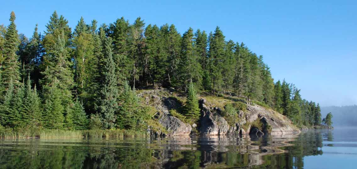 Still water reflects a rocky cliff covered in evergreen trees.