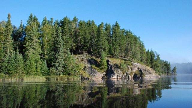 A large still body of water reflects a rocky shore and evergreen trees.