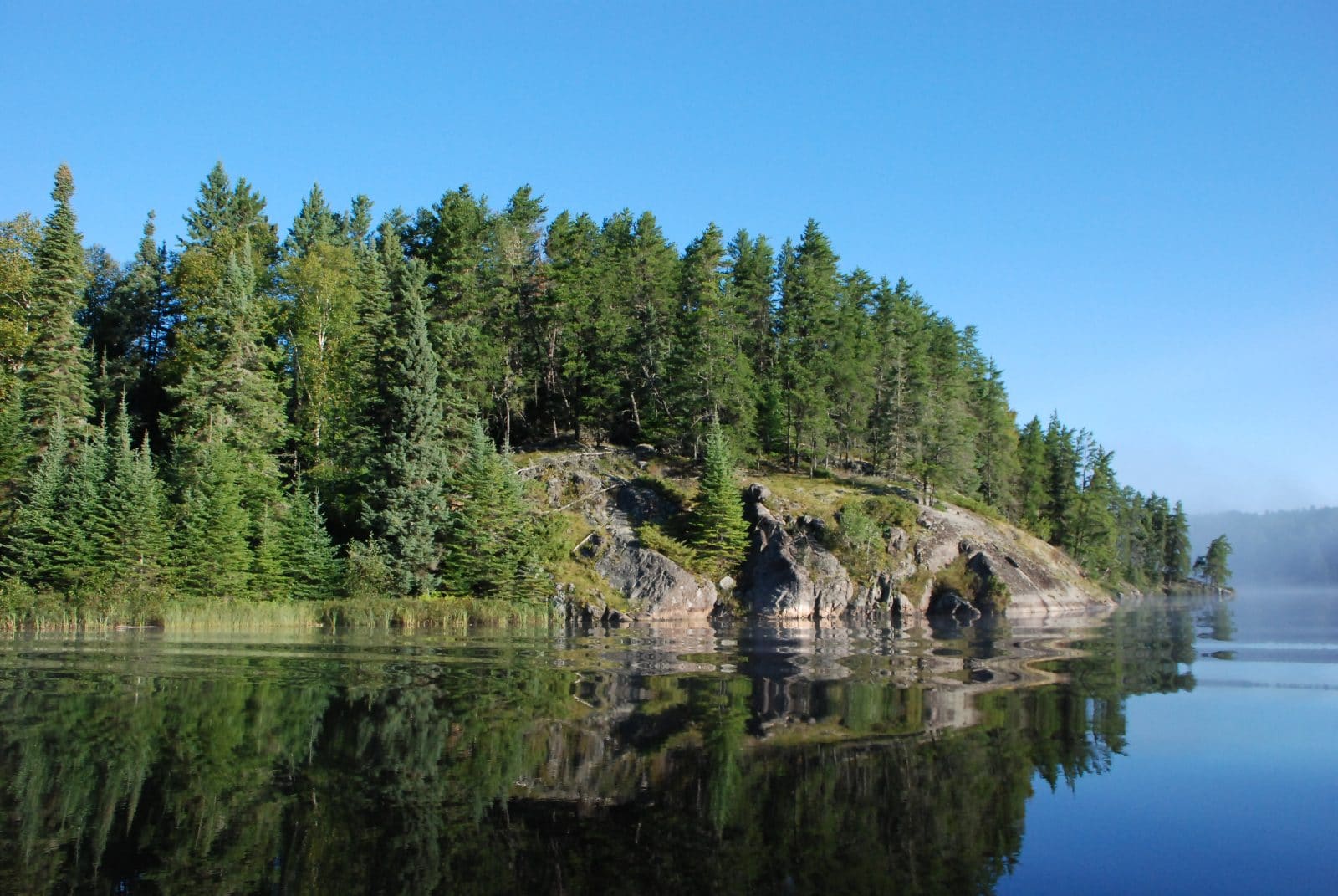 A large still body of water reflects a rocky shore and evergreen trees.
