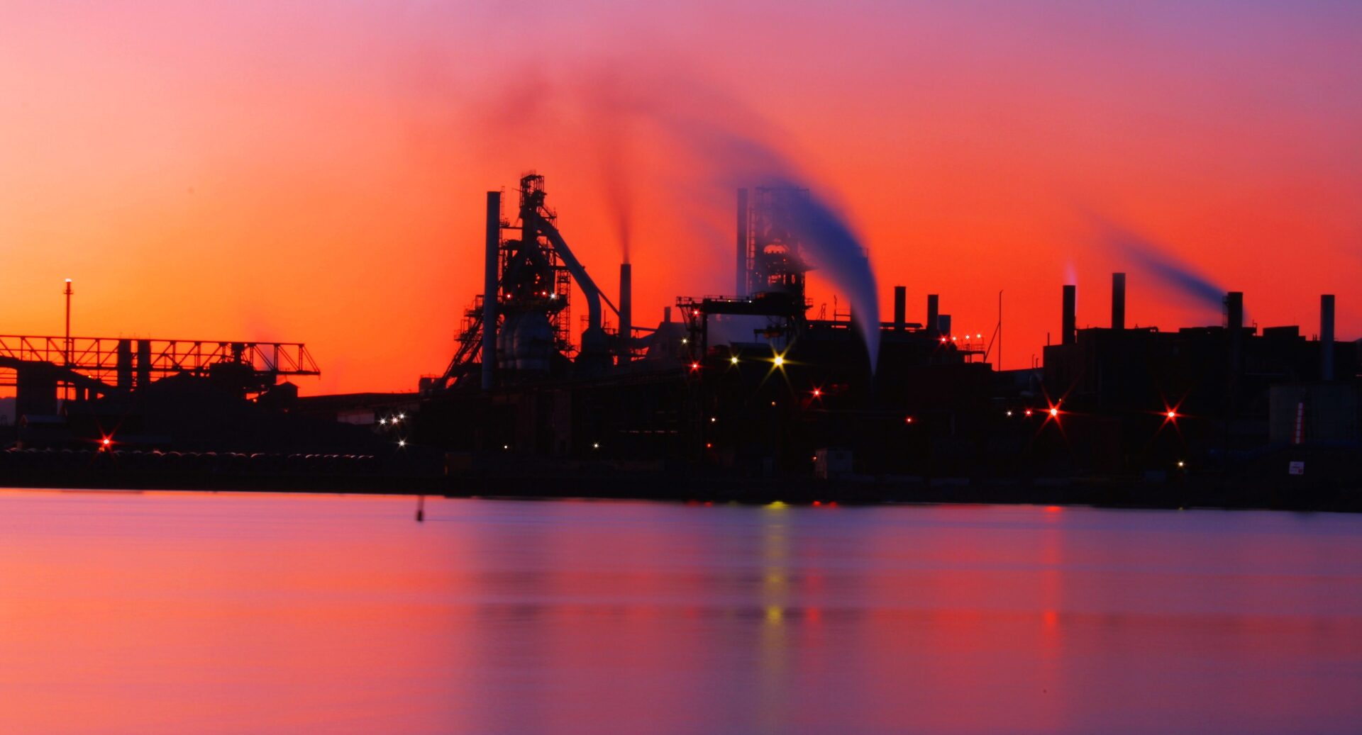An industrial complex at sunset. The complex is built near a body of water and large plumes of smoke rise from smoke stacks.