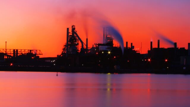 An industrial complex at sunset. The complex is built near a body of water and large plumes of smoke rise from smoke stacks.