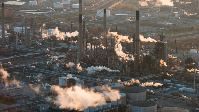 a large industrial complex with tall smoke stacks. Smoke rises from many different stacks and creates plumes of smoke in the air.