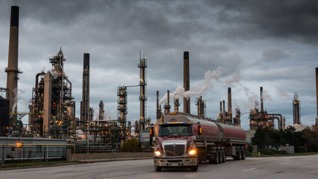 A large truck is parked outside of an industrial complex. Smoke billows out of large smoke stacks.