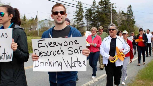 A crowd of people march down a road in Harrietsfield, NS. A man holds a sign reading 