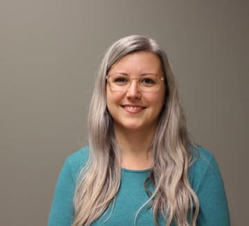 Suzie smiles at the camera. She has long silver hair and wears a light blue shirt and glasses. She stands against a grey background.