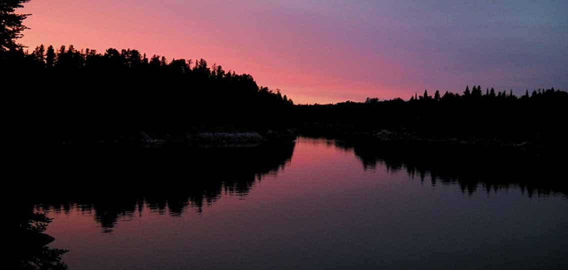 A still lake reflects trees on a shoreline and a soft pink sunset.