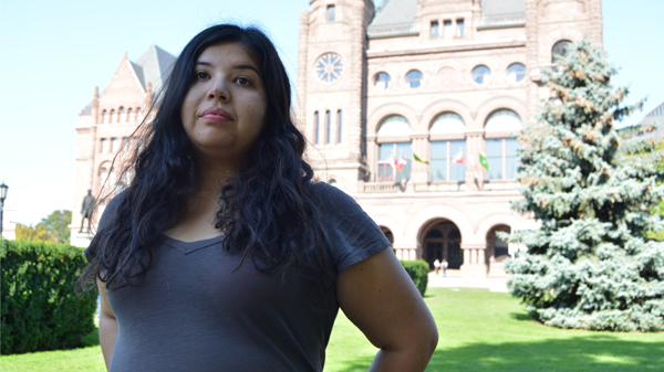 Vanessa stands in front of a parliament building. She has long wavy black hair and wears a grey t-shirt.