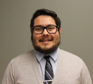 Jordano smiles at the camera. He wears a grey sweater and striped tie. He has short brown hair and brown facial hair. He wears square glasses.