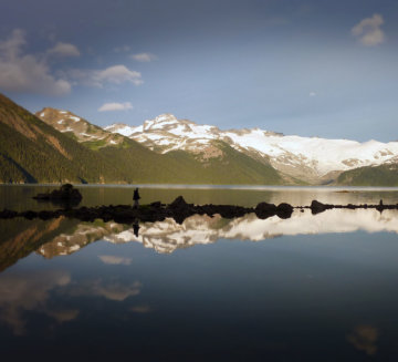 Snow-covered mountains stand against a blue sky. The mountains and sky are reflected in a still lake.