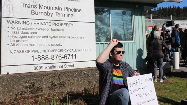A protester sits in front of the Trans Mountain Pipeline sign and holds a hand written sign reading
