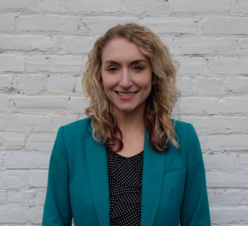 Danielle stands against a brickwork background. She wears a teal suit jacket and black blouse. She has curly, blonde hair.