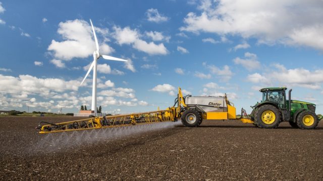 A tractor drives across a flat field spraying crops. A wind turbine stands in the background.