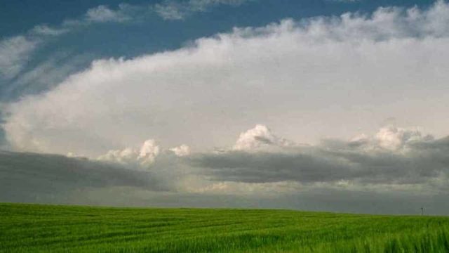 A flat, green field of grass is beneath a sky full of grey, stormy clouds.