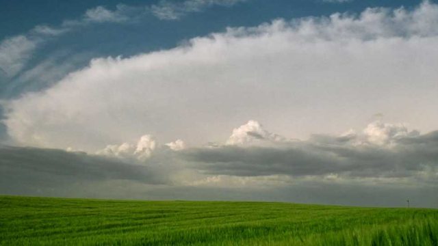 A flat grassy field has grey clouds that rise over the field.