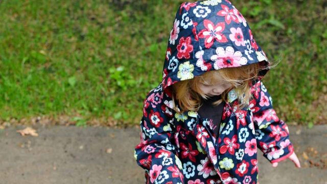 A toddler wears a floral rain coat and stands looking down in the mud.