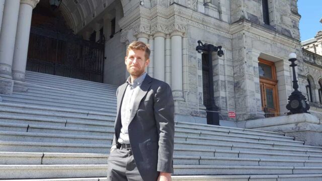 Alan stands outside of a parliament building. He smiles and wears a grey suit. He has light brown hair and facial hair.