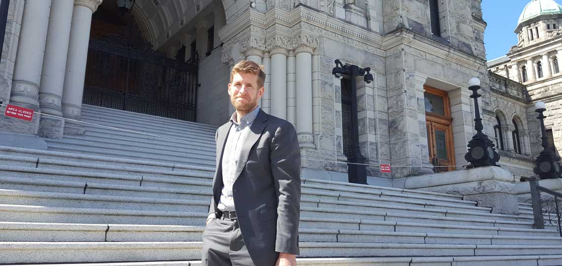 Alan stands outside of a parliament building. He smiles and wears a grey suit. He has light brown hair and facial hair.