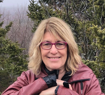 Deborah stands outside, in front of evergreen trees. She wears a pink jacket and pink glasses. She has shoulder-length blonde hair.