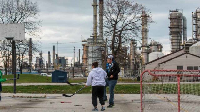 Youths play hockey in the street. An oil refinery appears in the background.