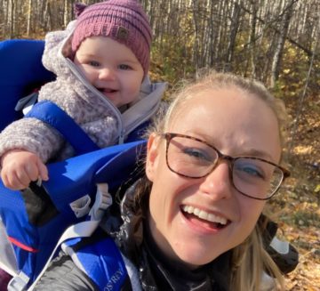 Jessica is outside and smiles at the camera. Her hair is in a ponytail and she wears square glasses. Her baby is in a carrier on her back and smiles wearing a pink toque.