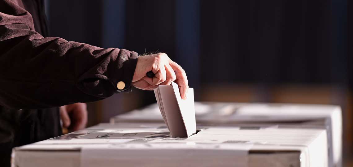 A hand places a folded ballot paper into a ballot box.