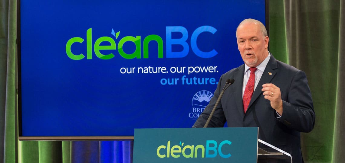Premier John Horgan speaks at a podium. The background logo reads 