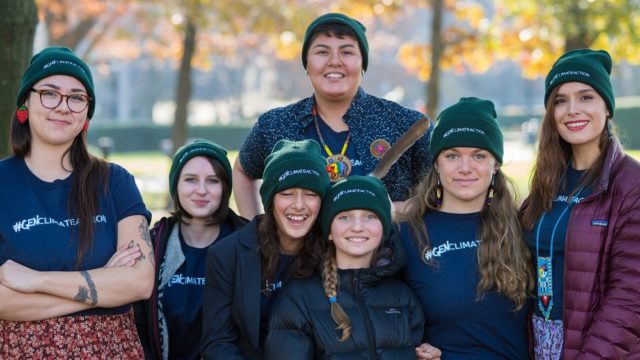 A group of youth stand together. They wear green toques and smile.