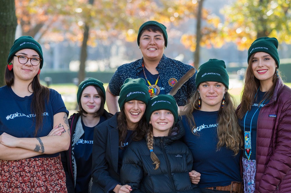 A group of youth stand together. They wear green toques and smile.