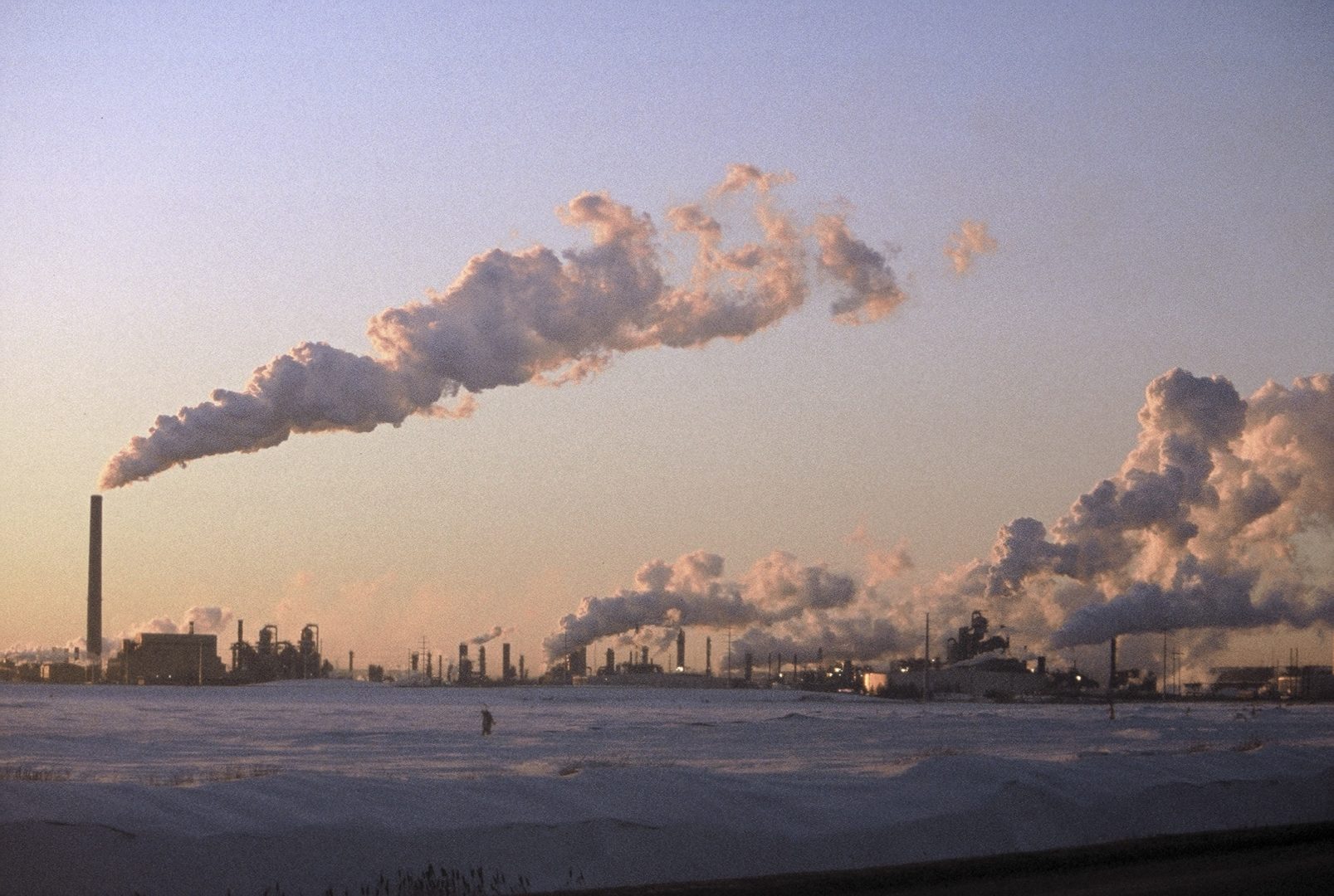 An industrial complex with many smoke stacks in the winter. Smoke rises from the stacks pouring into the sky.