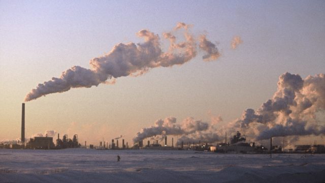 An industrial complex with many smoke stacks in the winter. Smoke rises from the stacks pouring into the sky.