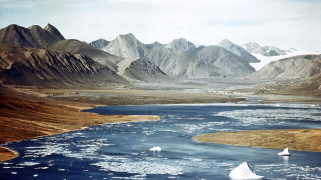 A blue river with broken ice flows toward tall mountains in the background.