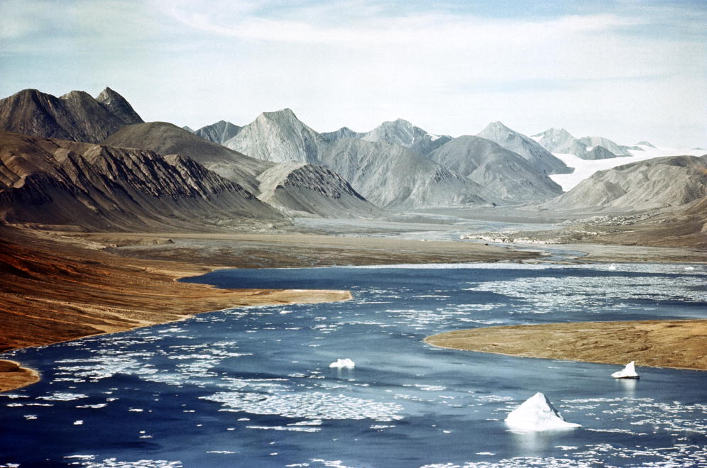 A blue river with broken ice flows toward tall mountains in the background.