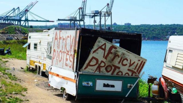 A trailer sits on the shore near water. On its side and back there are signs that read 