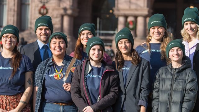 A group of 7 young people stand together and smile. They all wear green toques.