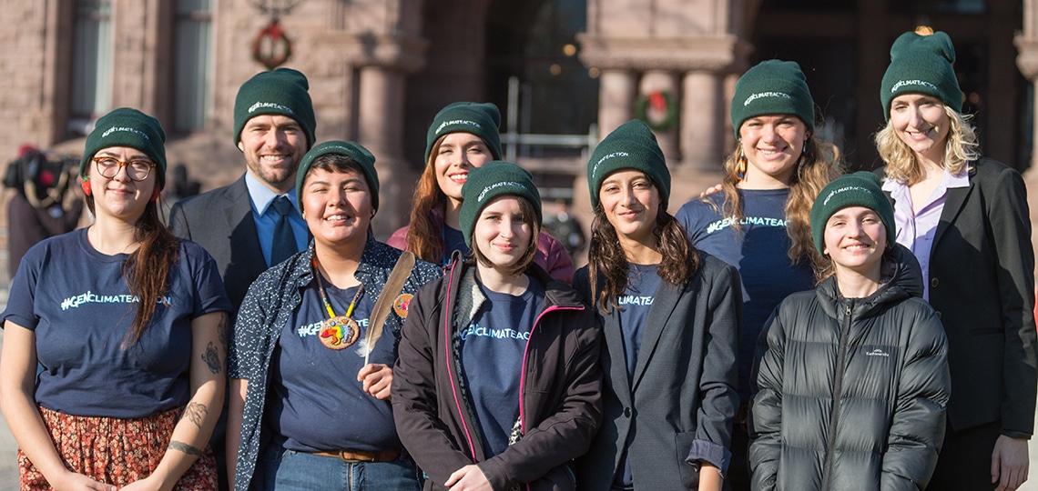 A group of 7 young people stand together and smile. They all wear green toques.
