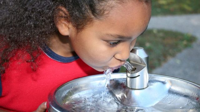 A young child drinks water from a silver water fountain as water flows from the spout.